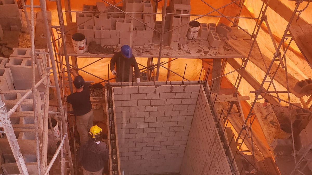 Scaffolding surrounds the construction zone, providing support and access to higher sections of the wall. The environment is typical of an active construction site, with workers focused on their tasks to erect a new building.