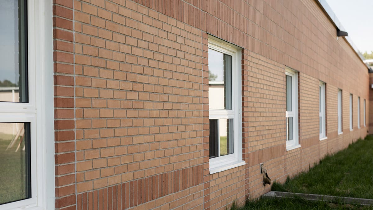 Side of a brick building with several windows. The bricks are arranged in a repeating pattern, and there is visible grass at the bottom of the wall. The windows have white frames and appear to be closed.