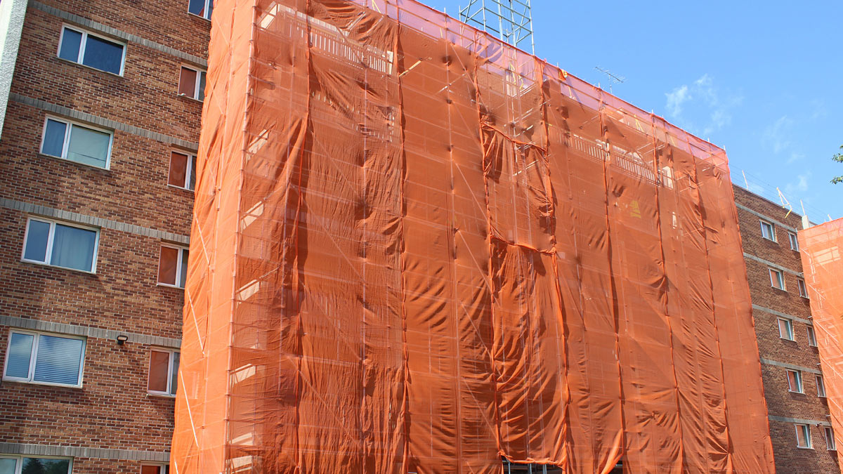 Multi-unit building under renovation, covered with orange safety netting and scaffolding for safe construction. The brick facade remains partially visible during the upgrade process.