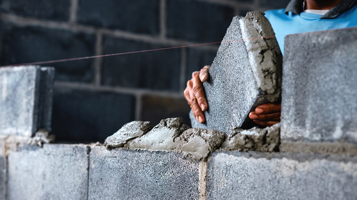 The image shows a close-up of a person’s hands placing a cinder block onto a mortar bed, constructing a wall. A string line is visible, used for alignment and levelness of the blocks. This image highlights the manual skill and precision involved in masonry work.