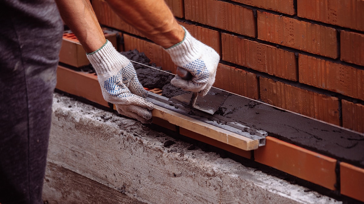 Worker wearing gloves applies mortar to a row of bricks, ensuring precise alignment and stability in a masonry project.